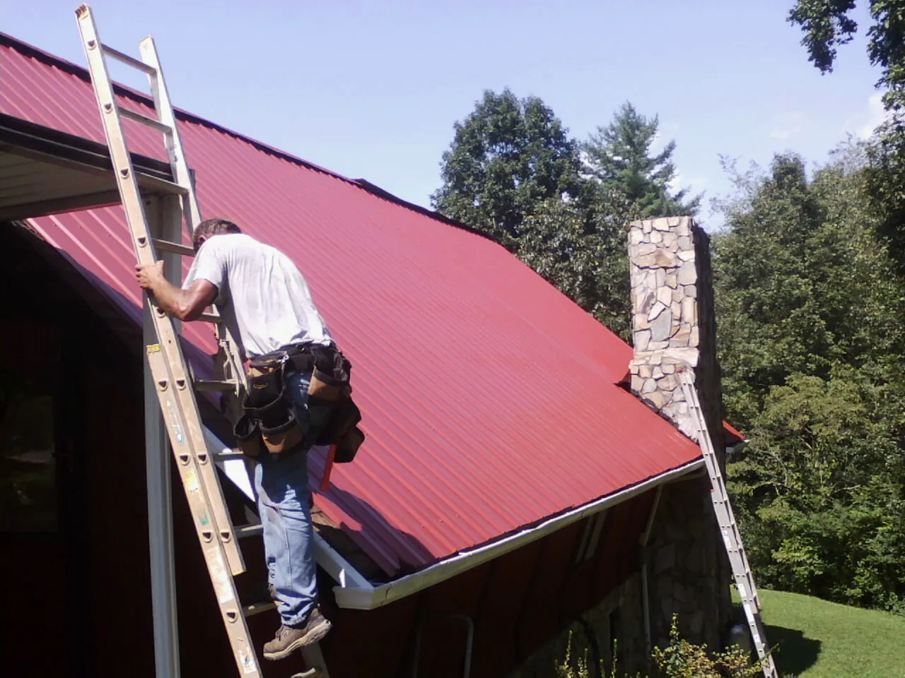 Roofer on a ladder beside a red metal roof and stone chimney — Soffit & Fascia Repair by Roofing Brothers in Indian Valley